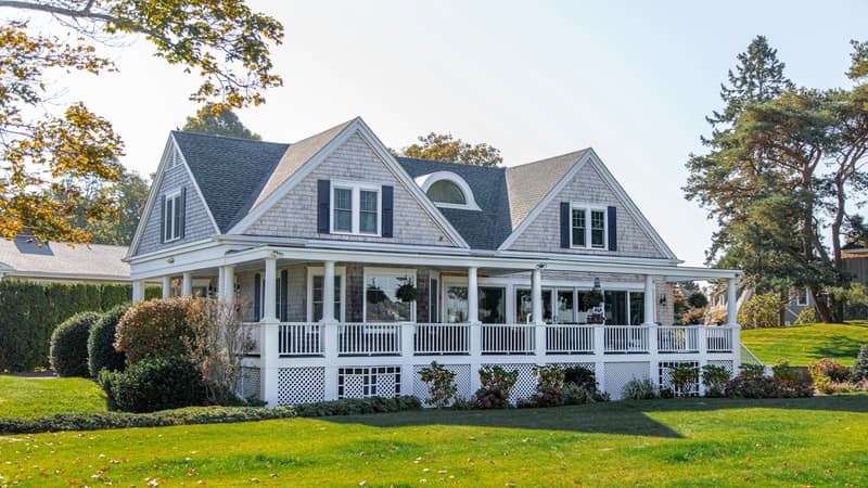 White shingled house with wraparound porch and manicured green lawn under clear sky